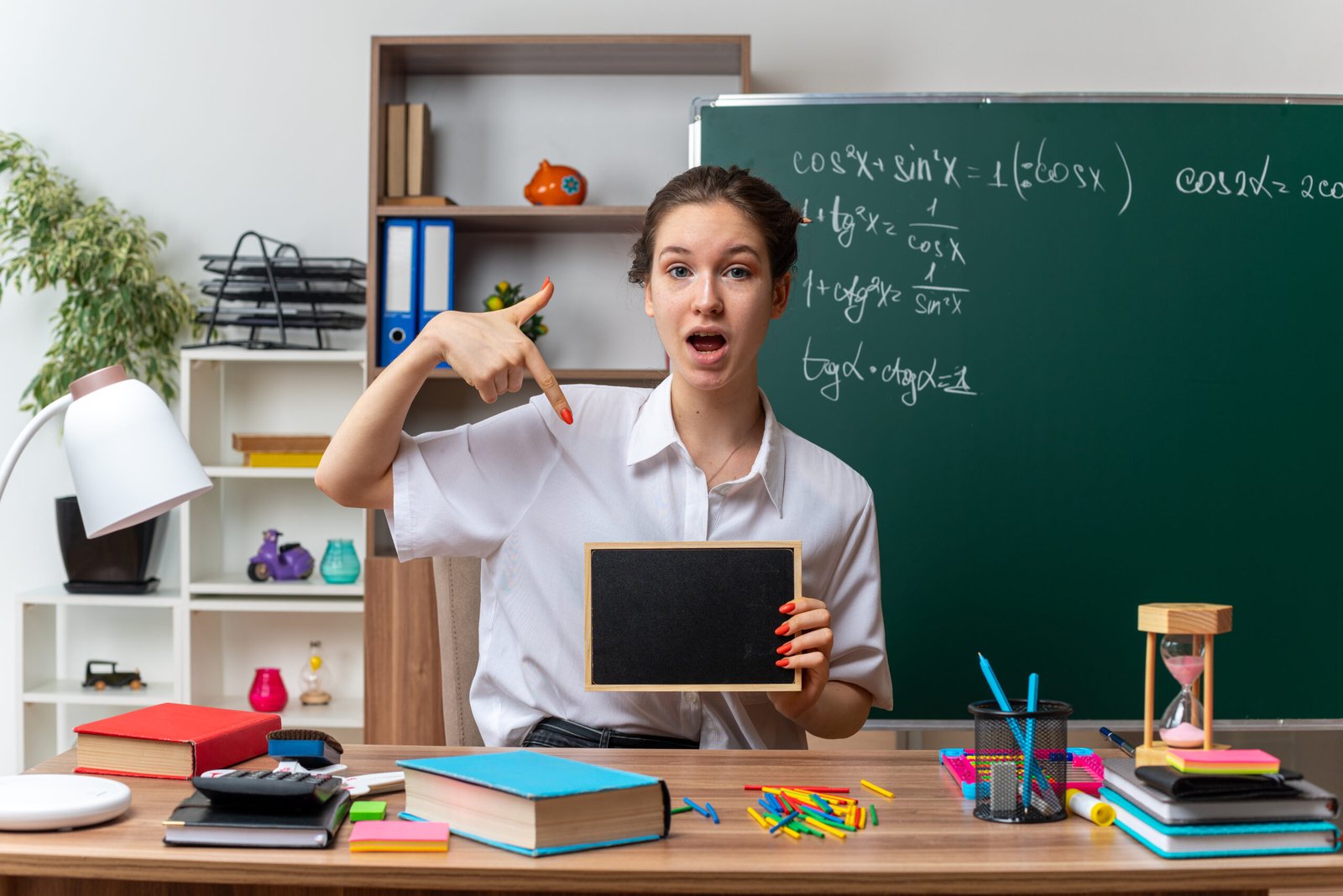 impressed young blonde female math teacher sitting desk with school tools holding pointing mini blackboard looking camera classroom